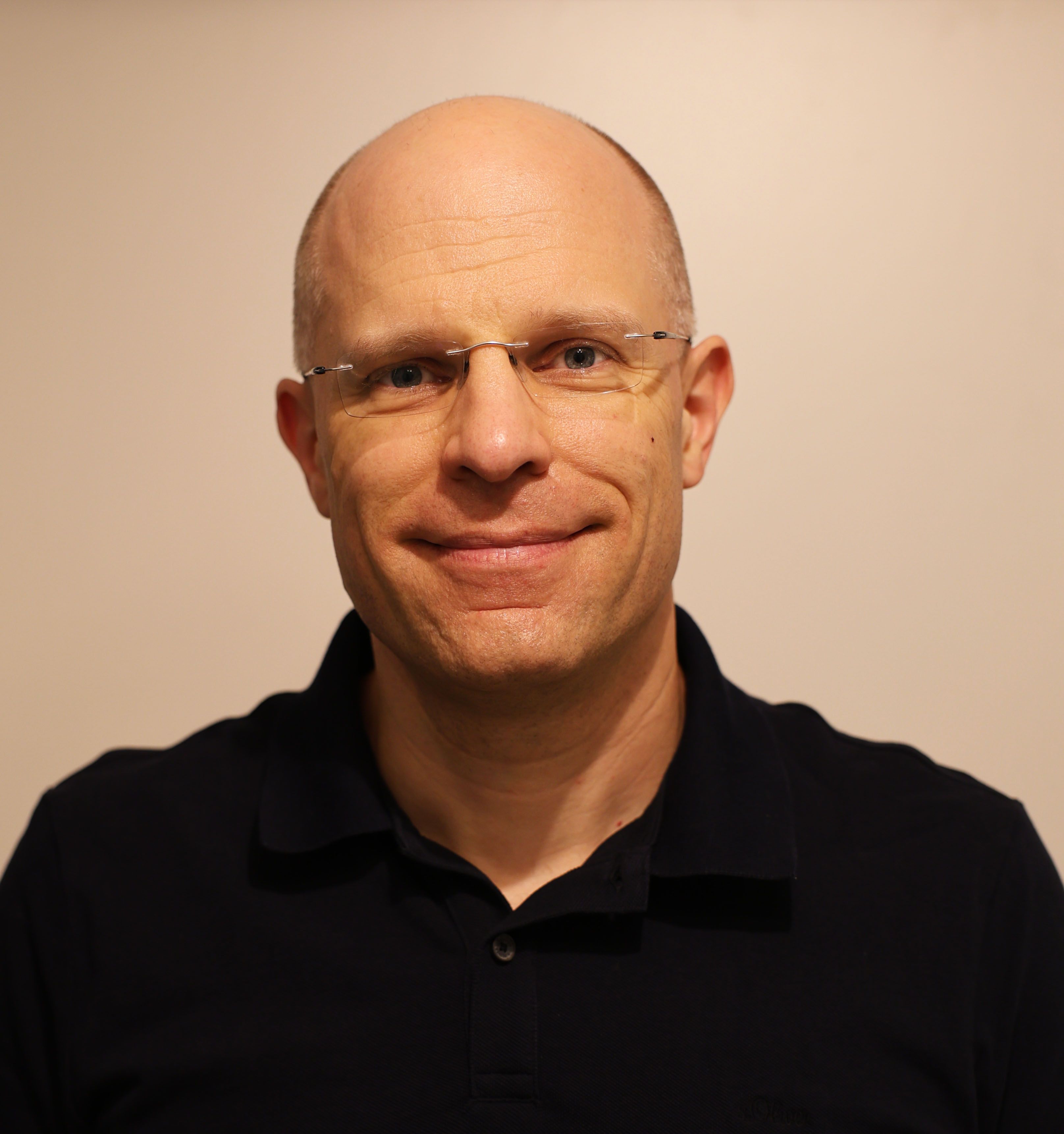 Portraitphoto eines Mannes mit Glatze und Brille vor beigem Hintergrund. Er trägt ein schwarzes Hemd mit leicht geöffnetem Kragen.
